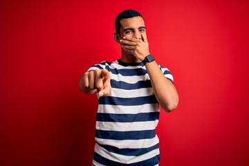 Fototapeta premium Handsome african american man wearing casual striped t-shirt standing over red background laughing at you, pointing finger to the camera with hand over mouth, shame expression