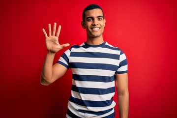 Naklejka premium Handsome african american man wearing casual striped t-shirt standing over red background showing and pointing up with fingers number five while smiling confident and happy.