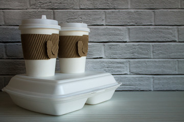 Two disposable coffee cups and a white container with food in close-up against a brick wall. The concept of food delivery takeaway