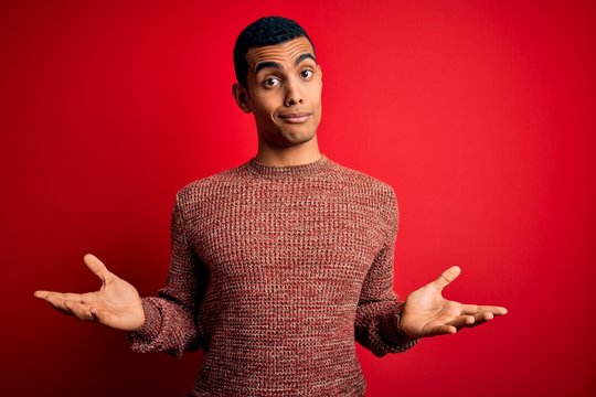 Young handsome african american man wearing casual sweater standing over red background clueless and confused expression with arms and hands raised. Doubt concept.
