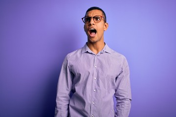 Obraz premium Handsome african american man wearing striped shirt and glasses over purple background angry and mad screaming frustrated and furious, shouting with anger. Rage and aggressive concept.