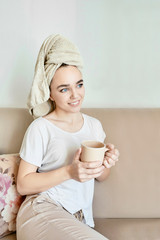 Young woman after a shower drinking from a coffee mug or tea sitting on a sofa. On the head is a towel after a shower.