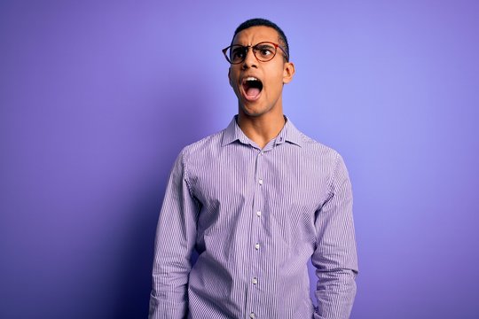 Handsome african american man wearing striped shirt and glasses over purple background angry and mad screaming frustrated and furious, shouting with anger. Rage and aggressive concept.