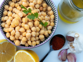 Chickpeas with Coriander leaves in a blue bowl, a lemon, a spoon with paprika, a head of garlic and a bottle of olive oil on white background