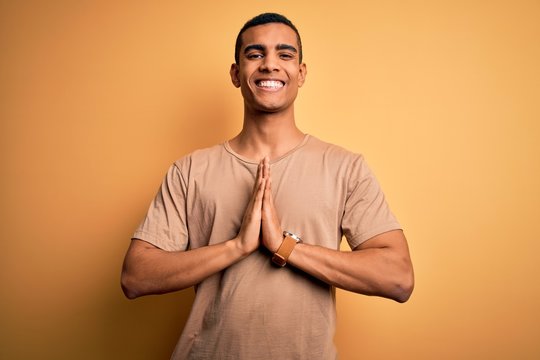 Young handsome african american man wearing casual t-shirt standing over yellow background praying with hands together asking for forgiveness smiling confident.