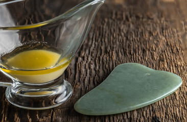 Jade scraper for gua cha and melted ghee in a glass cup on a background of wood texture.