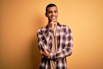 Fototapeta premium Young handsome african american man wearing casual shirt standing over yellow background looking confident at the camera smiling with crossed arms and hand raised on chin. Thinking positive.
