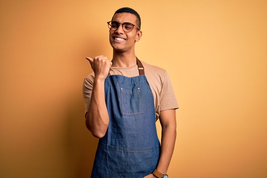 Young handsome african american shopkeeper man wearing apron over yellow background smiling with happy face looking and pointing to the side with thumb up. - Powered by Adobe