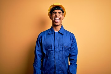 Young handsome african american worker man wearing blue uniform and security helmet sticking tongue out happy with funny expression. Emotion concept.