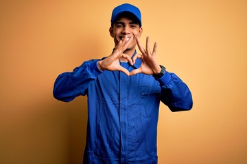 Young african american mechanic man wearing blue uniform and cap over yellow background smiling in love doing heart symbol shape with hands. Romantic concept.