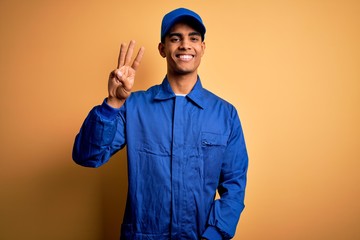 Young african american mechanic man wearing blue uniform and cap over yellow background showing and pointing up with fingers number three while smiling confident and happy.