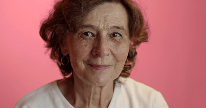 Senior woman against pink background looking straight at camera putting face mask off. Head and shoulders of old woman wearing protection mask. Hand-held shot, single shot, close up, 4K.