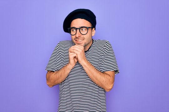 Handsome man with blue eyes wearing striped t-shirt and french beret over purple background laughing nervous and excited with hands on chin looking to the side