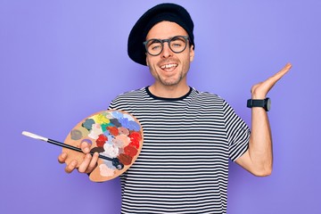 Young artist man with blue eyes wearing french beret and glasses drawing using paintbrush very happy and excited, winner expression celebrating victory screaming with big smile and raised hands