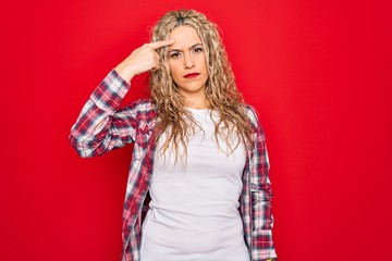 Young beautiful blonde woman wearing casual shirt standing over isolated red background pointing unhappy to pimple on forehead, ugly infection of blackhead. Acne and skin problem