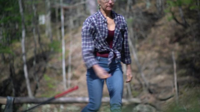 Side shot of mature woman wearing flannel and jeans in a forest trying to split wood, but misses and starts laughing.