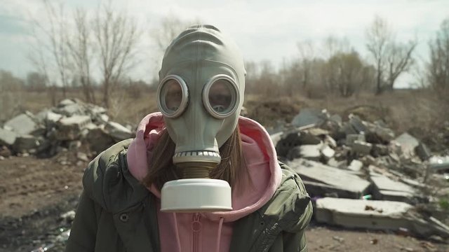 The girl in the gas mask turns to the camera and wears a pink hood. on the background dump