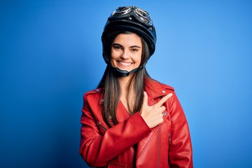 Young beautiful brunette motorcycliste woman wearing motorcycle helmet and jacket cheerful with a smile of face pointing with hand and finger up to the side with happy and natural expression on face
