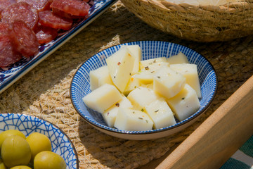 Close up of a blue plate filled of seasoned cheese with herbs and olive oil next to spanish tapas of olives and smoked sausage.