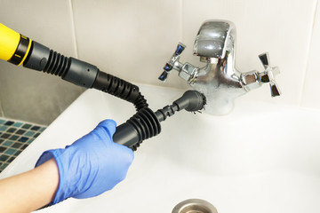 cleaning white bathroom sink and tap with yellow steam generator, hand in blue glove holds black...