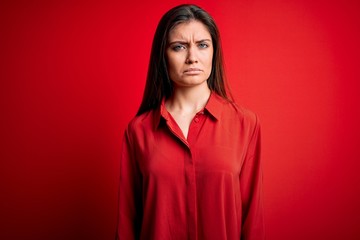 Young beautiful woman with blue eyes wearing casual shirt standing over red background skeptic and...