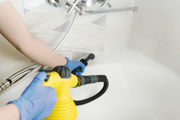 cleaning white bathtub and chrome tap with steam generator, hand in blue glove holds yellow steam...