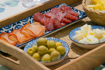 Close up of a wooden tray filled with spanish tapas with olives, cheese, smoked sausage, slices of ham and a bowl of crisps