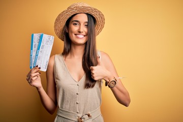 Young beautiful brunette tourist woman on vacation wearing hat holding boarding pass happy with big smile doing ok sign, thumb up with fingers, excellent sign