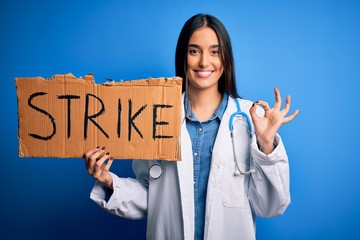 Young doctor woman wearing stethoscope holding cardboard banner protesting in strike doing ok sign with fingers, excellent symbol