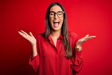 Young beautiful brunette woman wearing casual shirt and glasses over red background celebrating mad and crazy for success with arms raised and closed eyes screaming excited. Winner concept