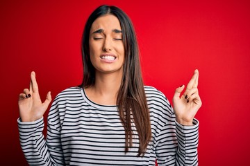 Young beautiful brunette woman wearing casual striped t-shirt over red background gesturing finger crossed smiling with hope and eyes closed. Luck and superstitious concept.