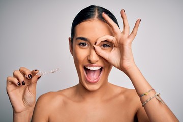 Young beautiful brunette woman holding dental aligner orthodontic to teeth corretion with happy face smiling doing ok sign with hand on eye looking through fingers