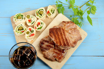two pieces of grilled meat on a cooking plate with pieces of tortilla next to a cold drink on an old blue table