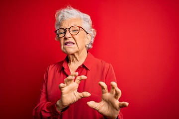 Senior beautiful grey-haired woman wearing casual shirt and glasses over red background disgusted expression, displeased and fearful doing disgust face because aversion reaction. With hands raised