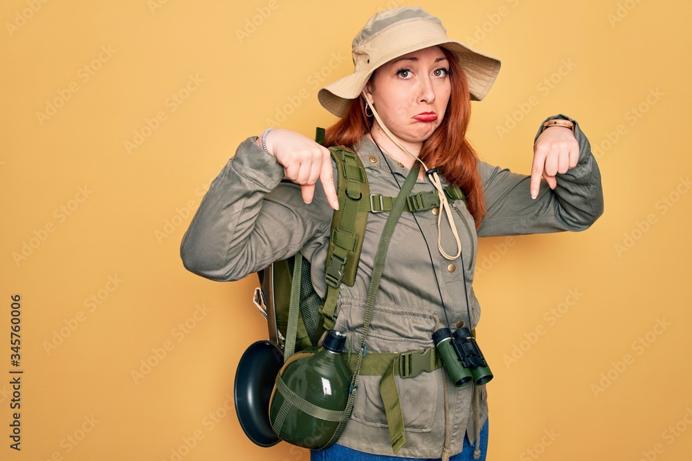 Canvas Prints young redhead backpacker woman hiking wearing backpack and hat over yellow background pointing down 