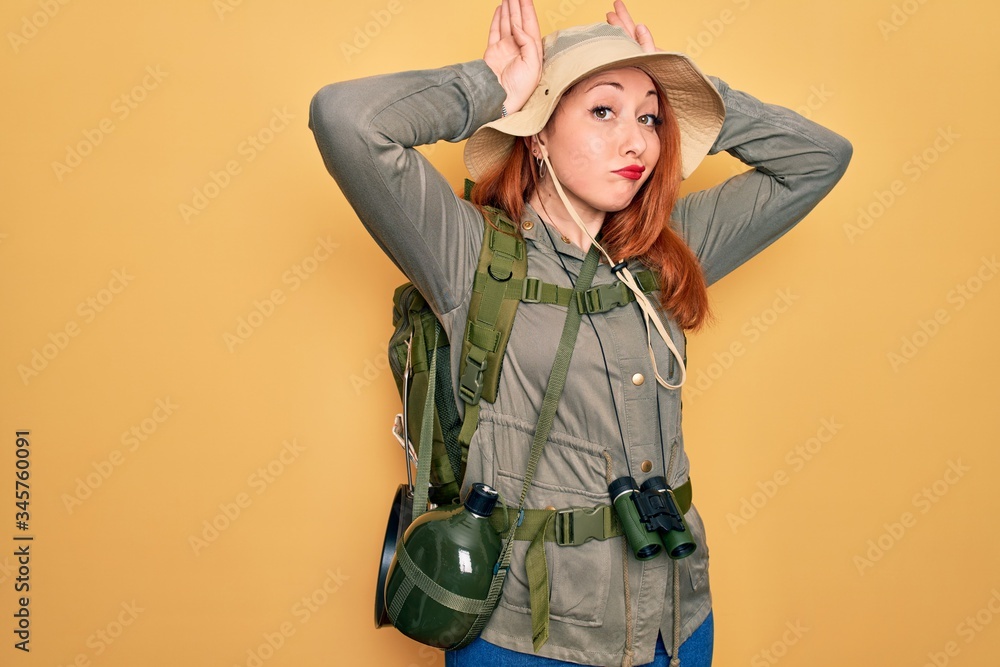 Wall mural Young redhead backpacker woman hiking wearing backpack and hat over yellow background Doing bunny ears gesture with hands palms looking cynical and skeptical. Easter rabbit concept.