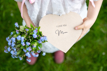 I love you mum written on a heart-shaped card and forget-me-nots held by a girl on grass background