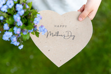 Happy Mother's Day written on a heart-shaped card and forget me nots held by a girl on grass background
