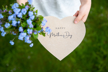 Happy Mother's Day written on a heart-shaped card and forget me nots held by a girl on grass background