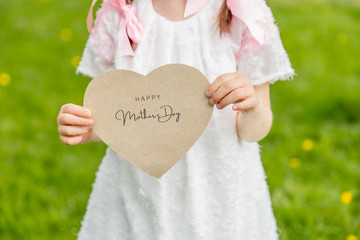 Happy Mother's Day written on a heart-shaped card held by a girl on grass background