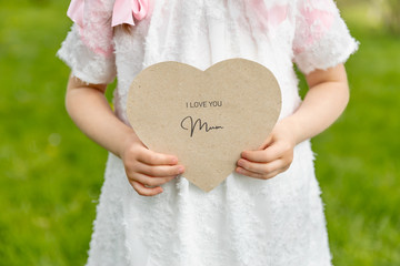 I love you mum written on a heart-shaped card held by a girl on grass background