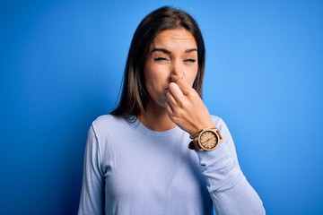 Young beautiful brunette woman wearing casual sweater standing over blue background smelling...