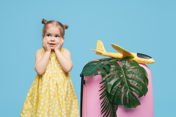 Cute baby girl in a summer dress with a pink suitcase is waiting for travel to tropical countries after quarantine. Blue studio background. Funny face