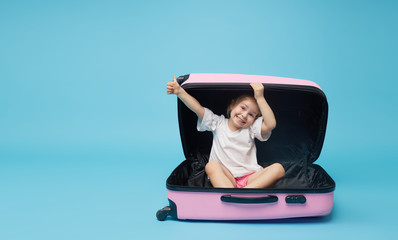 Cute baby girl sitting in a suitcase and waiting for summer vacation after quarantine. Pointing to the side. Blue studio background.