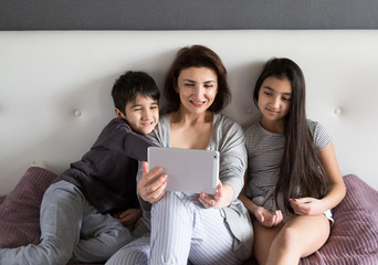 Mother and small kids having happy family time at home watching movie on tablet, taking a selfie, enjoying domestic life together.