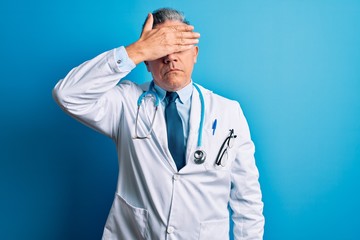 Middle age handsome grey-haired doctor man wearing coat and blue stethoscope covering eyes with hand, looking serious and sad. Sightless, hiding and rejection concept