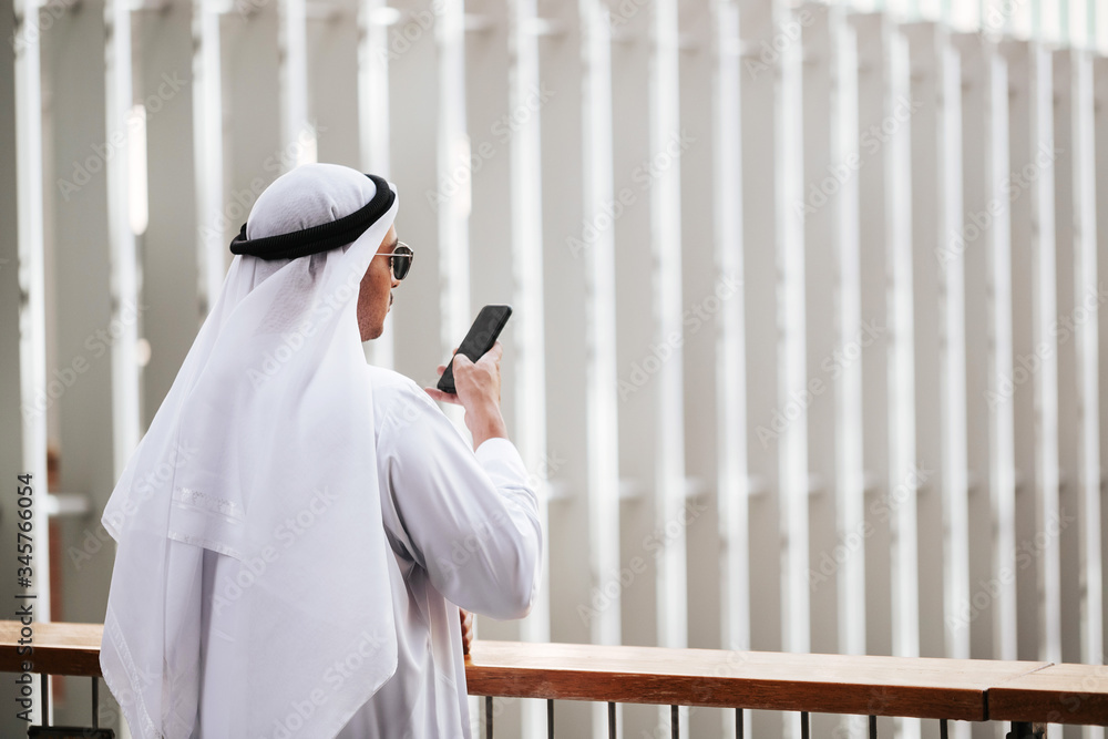 Wall mural Emirati guy wearing traditional kandura in urban city emirates lifestyle talking to smart phone. Side view photography concept for Arab magazine.