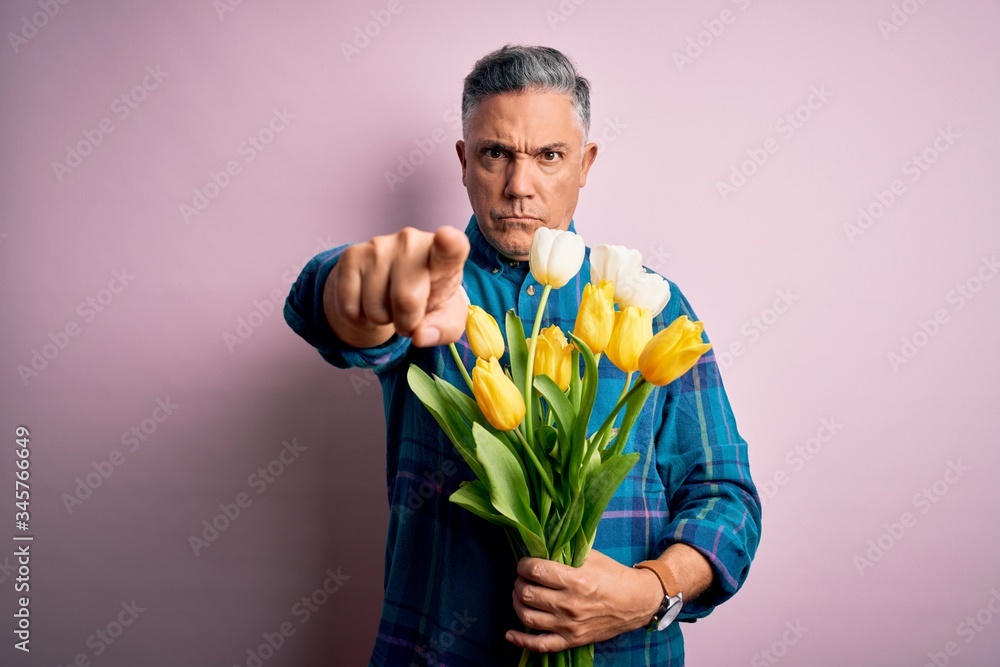 Poster Middle age grey-haired man holding natural bouquet of yellow tulips over pink background pointing with finger to the camera and to you, hand sign, positive and confident gesture from the front