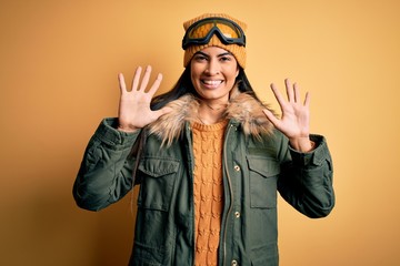 Young beautiful hispanic woman wearing ski glasses and coat for winter weather showing and pointing up with fingers number ten while smiling confident and happy.