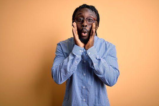 Young handsome african american man wearing shirt and glasses over yellow background afraid and shocked, surprise and amazed expression with hands on face